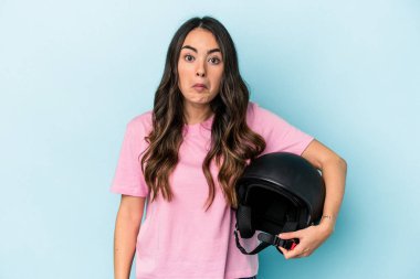 Young caucasian woman holding a motor bike helmet isolated on blue background shrugs shoulders and open eyes confused.