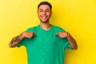 Sarı arka planda izole edilmiş beyaz dövmeli genç bir adam parmağıyla işaret ederek şaşırdı, geniş bir gülümsemeyle..
