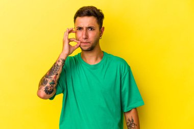 Sarı arka planda dövmeleri olan genç beyaz bir adam. Parmakları dudaklarında bir sır saklıyor..