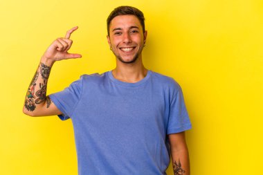 Sarı arka planda izole edilmiş beyaz dövmeli genç bir adam işaret parmaklarıyla küçük bir şey tutuyor, gülümsüyor ve kendinden emin..