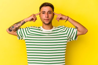 Sarı arka planda izole edilmiş dövmeleri olan beyaz genç bir adam görevine odaklanmış, işaret parmaklarını işaret ediyor..