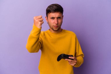 Mor arka planda izole edilmiş oyun kumandası tutan genç beyaz adam kameraya yumruk gösteriyor, agresif yüz ifadesi..