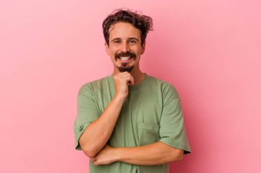Pembe arka planda izole edilmiş genç beyaz adam gülümsüyor ve kendinden emin, elleriyle çenesine dokunuyor..