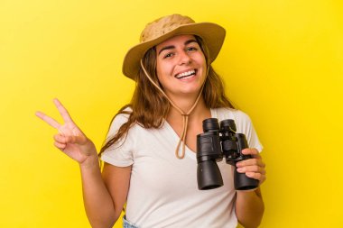 Sarı arka planda dürbün tutan genç beyaz kadın neşeli ve kaygısız parmaklarıyla barış sembolü gösteriyor..