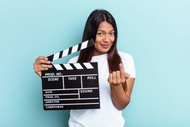 Genç Venezuelalı bir kadın mavi arka planda izole edilmiş bir klempon tutuyor. Sanki seni davet ediyormuş gibi parmakla gösteriyor..