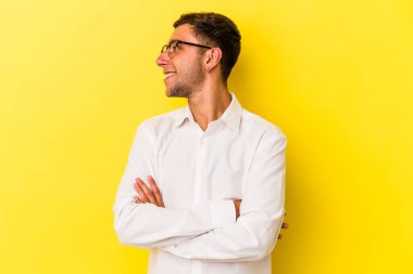 Sarı arka planda izole edilmiş dövmeleri olan genç beyaz adam kollarını kavuşturarak gülümsüyor..