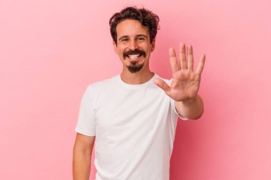 Pembe arka planda izole edilmiş genç beyaz adam gülümseyerek beş numarayı parmaklarıyla gösteriyor..