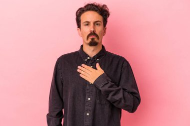 Pembe arka planda izole edilmiş genç beyaz adam yemin ediyor, elini göğsüne koyuyor..