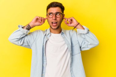 Sarı arka planda dövmeleri olan, kulaklarını parmaklarıyla kapatan, stresli ve çaresiz genç beyaz bir adam..