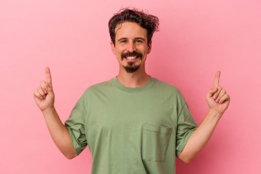 Pembe arka planda izole edilmiş genç beyaz adam iki parmağı da boş bir alan gösteriyor..