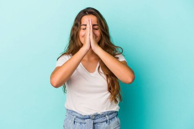 Mavi arka planda izole edilmiş genç beyaz bir kadın el ele tutuşup ağza yakın dua ediyor, kendinden emin..