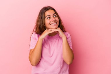 Pembe arka planda izole edilmiş genç beyaz kadın ellerini çenesinin altında tutuyor, mutlu bir şekilde yana bakıyor..
