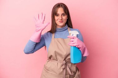 Pembe arka planda sprey tutan beyaz tenli genç kadın gülümseyerek beş numarayı parmaklarıyla gösteriyor..