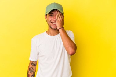 Sarı arka planda dövmeleri olan beyaz bir adam yüzünün yarısını avuç içiyle kaplayarak eğleniyor..