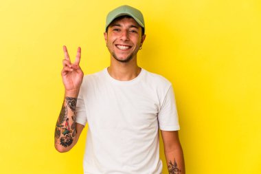 Sarı arka planda izole edilmiş dövmeleri olan genç beyaz adam zafer işareti gösteriyor ve gülümsüyor..