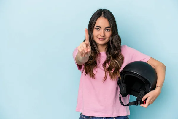Young caucasian woman holding a motor bike helmet isolated on blue background showing number one with finger.