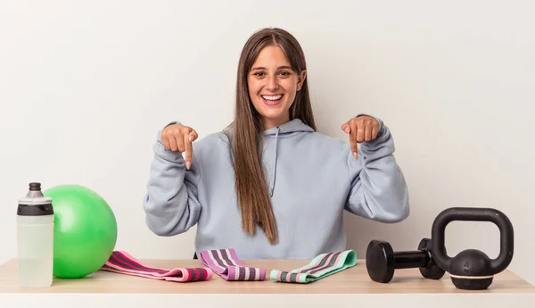 Young caucasian woman sitting at a table with sport equipment isolated on white background points down with fingers, positive feeling.
