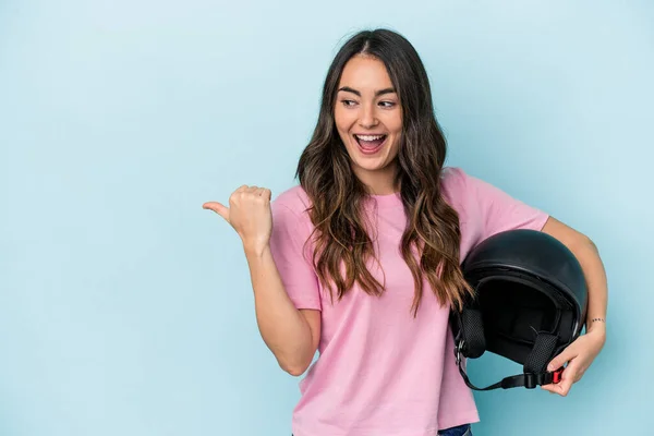 Young caucasian woman holding a motor bike helmet isolated on blue background points with thumb finger away, laughing and carefree.
