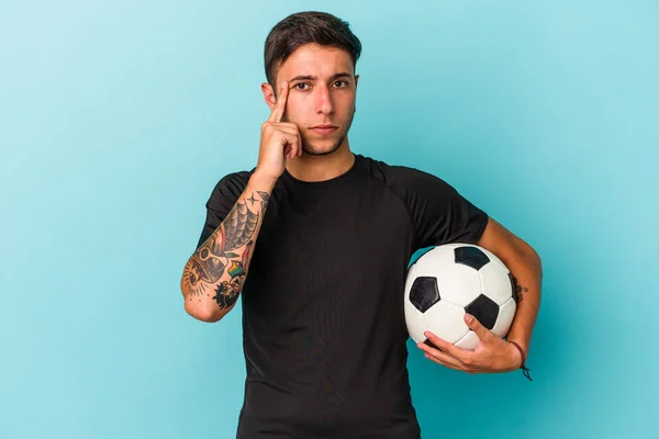 Młody Człowiek Gra Piłkę Nożną Trzymając Piłkę Niebieskim Tle Wskazując — Zdjęcie stockowe