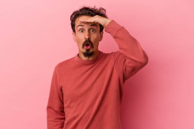 Pembe arka planda izole edilmiş genç beyaz adam uzaklara bakıyor ve elini alnına koyuyor..