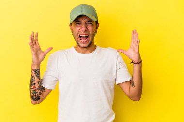 Sarı arka planda izole edilmiş dövmeleri olan genç beyaz adam gökyüzüne haykırıyor, yukarı bakıyor, hayal kırıklığına uğruyor..