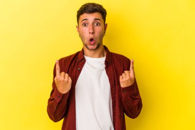 Sarı arka planda izole edilmiş, ağzı açık, üst tarafı işaret eden dövmeleri olan beyaz bir genç..