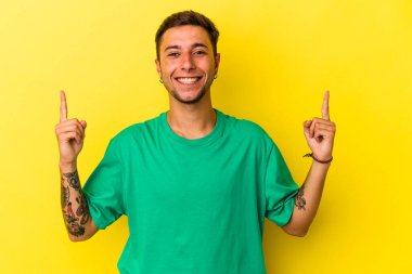 Sarı arka planda dövmeleri olan beyaz genç bir adam iki ön parmağı da boş bir alan gösteriyor..