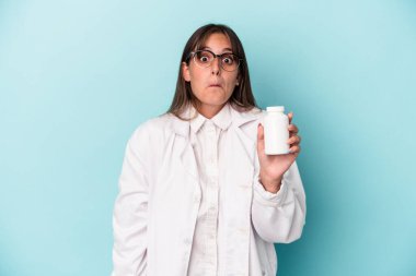 Mavi arka planda izole edilmiş haplar tutan genç eczacı kadın omuz silker ve gözleri açık kalır..