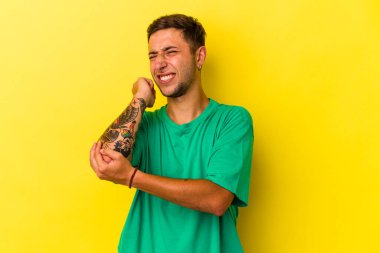 Dövmeleri olan beyaz bir genç adam sarı arka planda izole edilmiş dirsek masajı yapıyor, kötü bir hareketin ardından acı çekiyor..