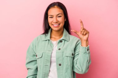 Pembe arka planda izole edilmiş genç Latin kadın işaret parmaklarıyla küçük bir şey tutuyor, gülümsüyor ve kendinden emin..