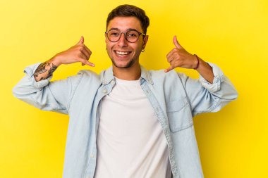 Sarı arka planda izole edilmiş dövmeleri olan genç beyaz bir adam parmakları olan bir cep telefonu hareketi gösteriyor..