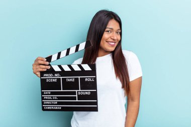 Genç Venezuelalı bir kadın mavi arka planda izole edilmiş bir el çırpma tahtası tutuyor. Gülümsüyor, neşeli ve keyifli görünüyor..