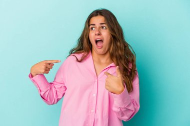 Mavi arka planda izole edilmiş genç beyaz kadın parmağını göstererek şaşırdı, geniş bir gülümsemeyle..