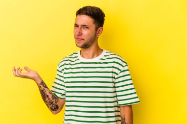 Sarı arka planda izole edilmiş dövmeleri olan genç, beyaz bir adam sorgulama sırasında omuz silkiyor ve şüphe duyuyor..