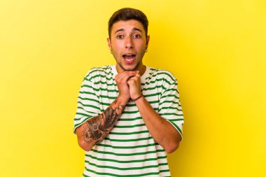 Sarı arka planda izole edilmiş beyaz dövmeli genç bir adam şans için dua ediyor, şaşırıyor ve ağzını açıyor..