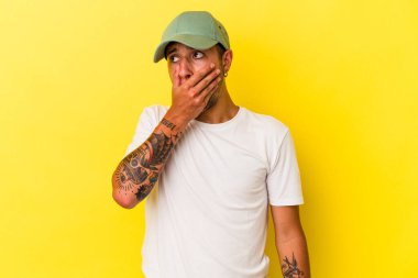 Sarı arka planda dövmeleri olan beyaz bir genç adam. Ağzını eliyle kapatan bir fotokopi odasına bakıyor..