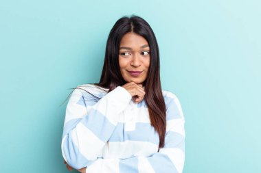 Genç Venezuelalı kadın mavi arka planda izole edilmiş şüpheli ve şüpheci bir ifadeyle yana bakıyor..