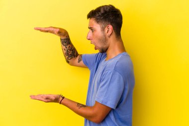 Sarı arka planda izole edilmiş beyaz dövmeli genç bir adam elleri arasında bir kopya alanı tutarken şok ve hayrete düştü..