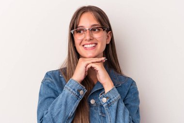 Beyaz arka planda izole edilmiş genç beyaz kadın ellerini çenesinin altında tutuyor, mutlu bir şekilde yana bakıyor..