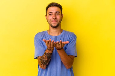 Sarı arka planda izole edilmiş beyaz dövmeli genç bir adam avuçlarıyla bir şey tutuyor ve kameraya sunuyor..