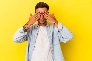 Sarı arka planda izole edilmiş dövmeleri olan genç beyaz bir adam elleriyle gözlerini kapatmaktan korkuyor..