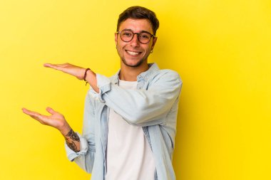 Sarı arka planda izole edilmiş beyaz dövmeli genç bir adam işaret parmaklarıyla küçük bir şey tutuyor, gülümsüyor ve kendinden emin..