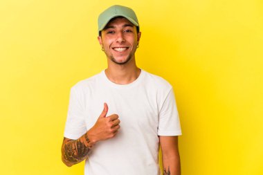 Sarı arka planda izole edilmiş dövmeleri olan genç beyaz adam gülümsüyor ve başparmağını kaldırıyor.