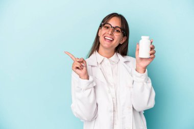 Mavi arka planda hapları saklayan genç bir eczacı kadın gülümsüyor ve boş alanda bir şeyler gösteriyor..