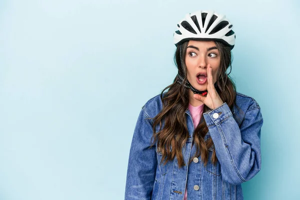 Young caucasian woman rinding a bike isolated on blue background is saying a secret hot braking news and looking aside