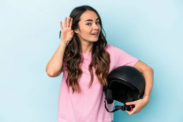 Young caucasian woman holding a motor bike helmet isolated on blue background trying to listening a gossip.