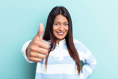 Genç Venezuelalı kadın mavi arka planda izole edilmiş gülümsüyor ve başparmağını kaldırıyor.