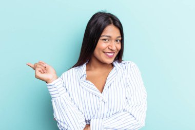 Genç Venezuelalı bir kadın mavi arka planda izole edilmiş, mutlu bir şekilde işaret parmağıyla işaret ederek gülümsüyor..