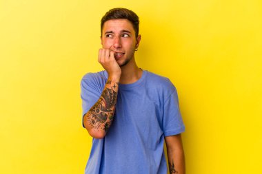 Sarı arka planda izole edilmiş, tırnak yiyen, gergin ve endişeli dövmeleri olan genç beyaz bir adam..