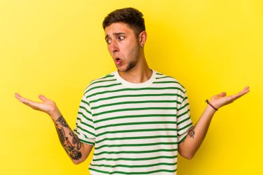Sarı arka planda izole edilmiş beyaz dövmeli genç bir adam. Kafası karışmış ve omuz silkiyor..